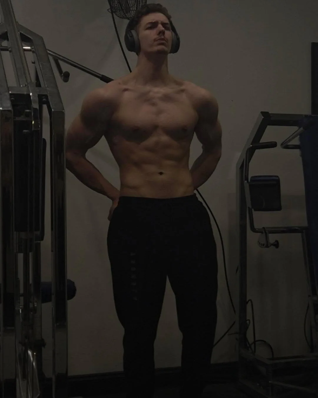Sam standing shirtless in the gym, headphones on, with a more developed physique